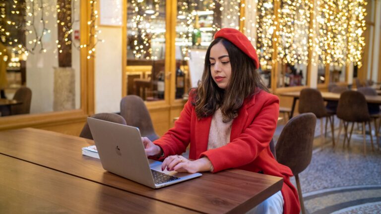 Young woman typing on laptop, wearing a red beret and coat, in a Parisian café.