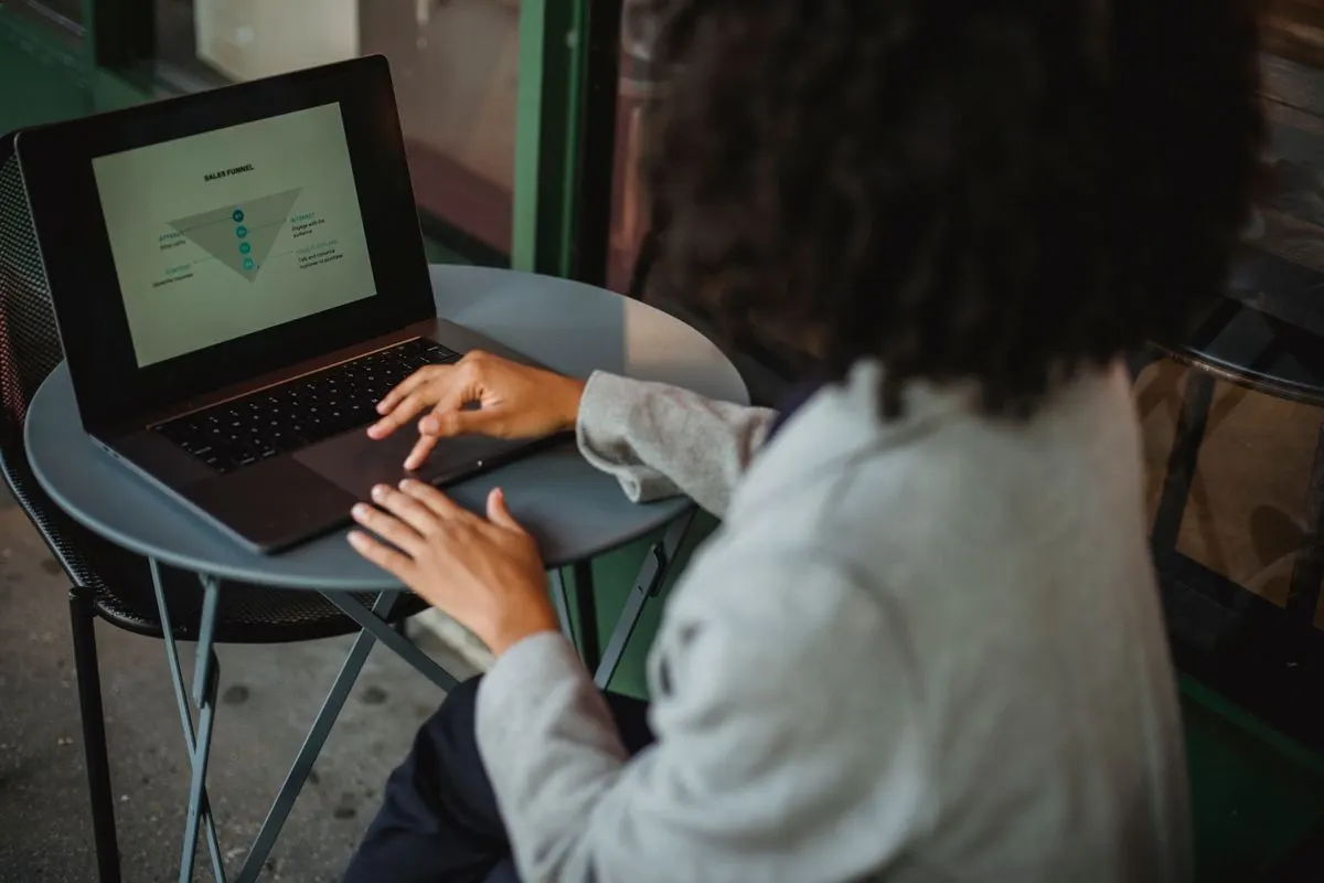 Back view of anonymous ethnic female in suit working with chart on computer while sitting at table in outdoor cafe during remote work